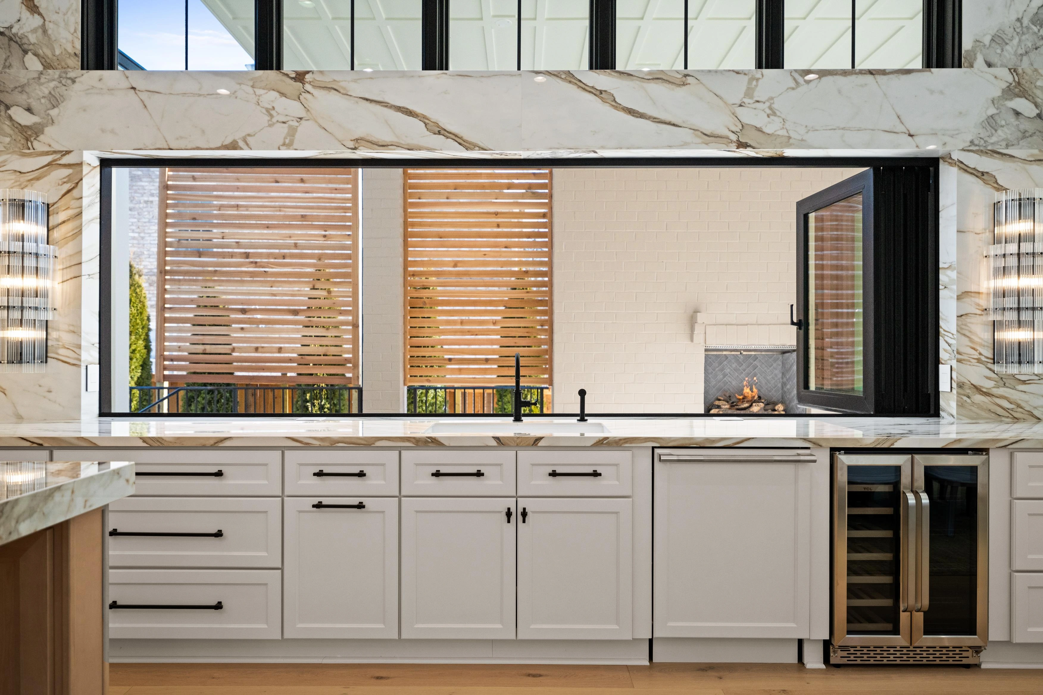 Kitchen pass-through window with calacatta marble surround, wine fridge, and view to covered patio with outdoor fireplace