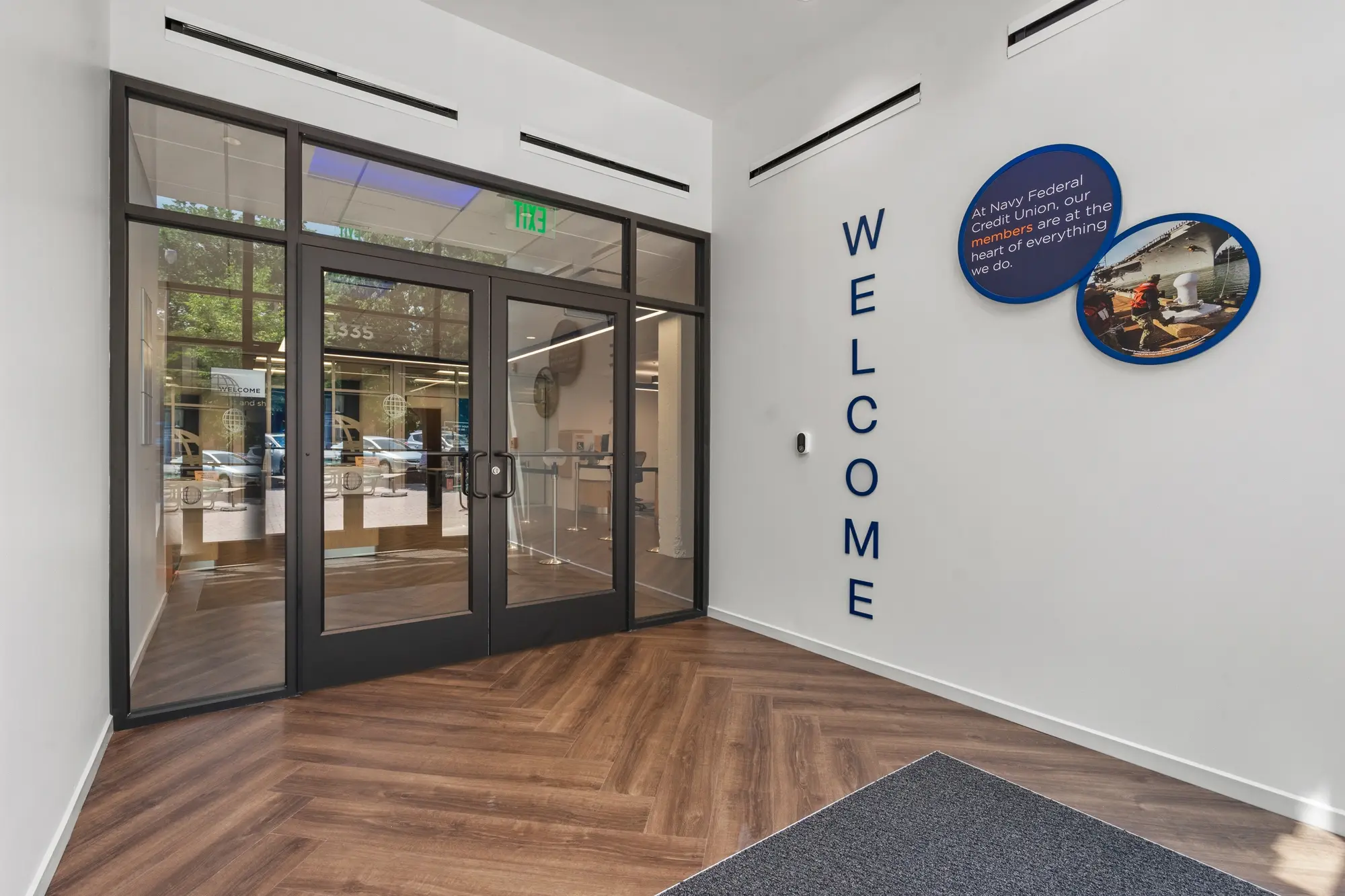 Branch foyer with herringbone vinyl plank flooring, vertical WELCOME lettering, and branded circular wall art