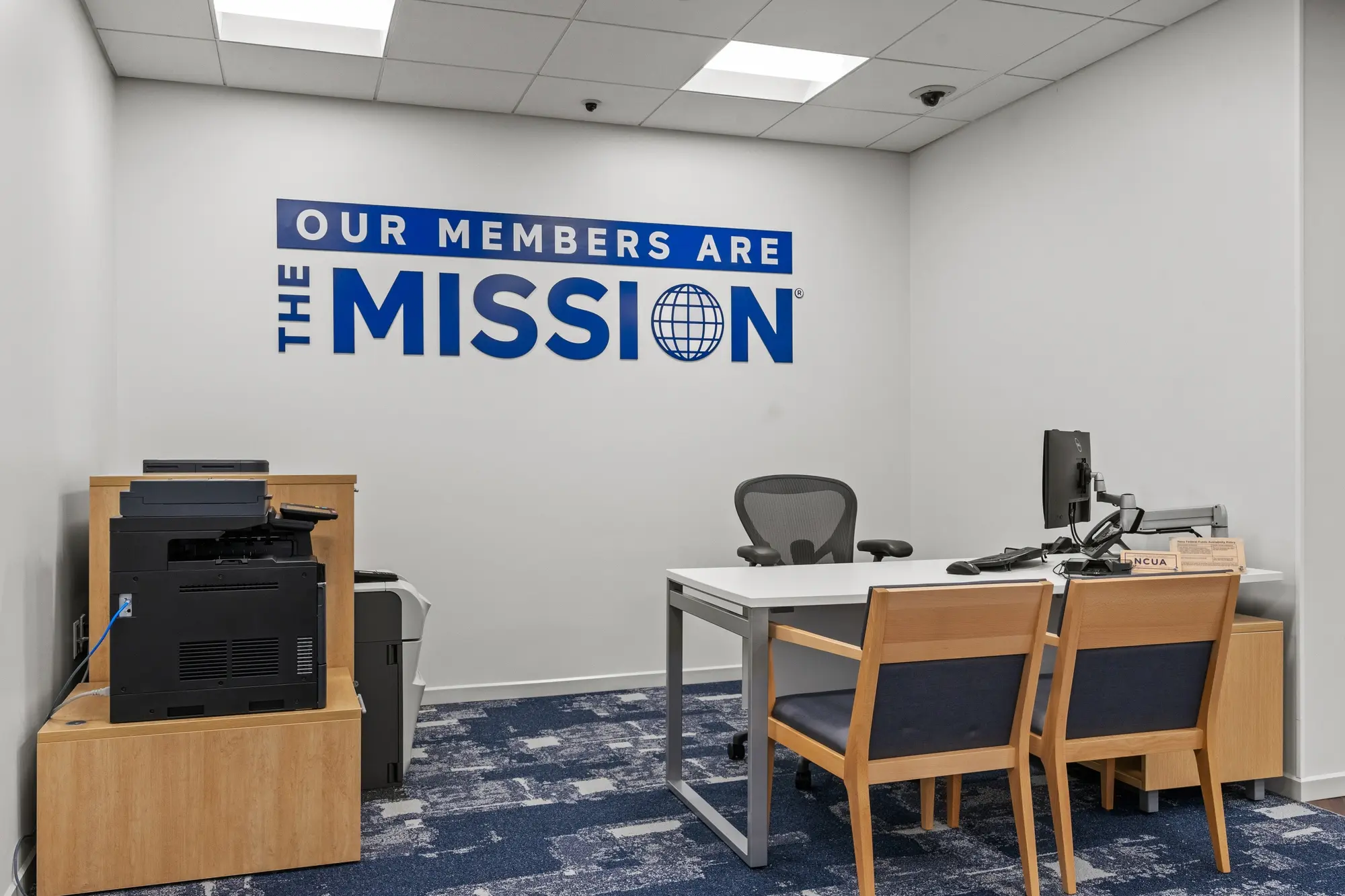 Advisor office with "Our Members Are The Mission" wall graphic, desk with dual monitors, printer station, and blue carpet